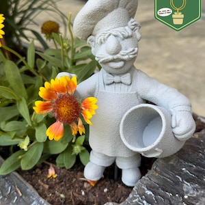 May include: A gray stone-textured garden figurine of a chef wearing a tall hat, bowtie, and apron. The chef holds a small hollow pot in one hand and a bright orange and yellow flower in the other. The figure has a mustache and textured surface.