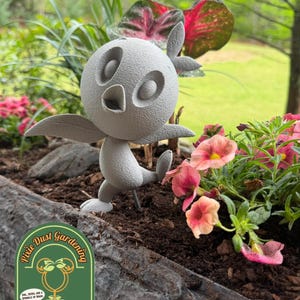 May include: A gray, whimsical bird-like garden statue with outstretched wings and a single leg raised, set in a planter with pink and yellow flowers. The planter has the text "Pixie Dust Gardening" on it.