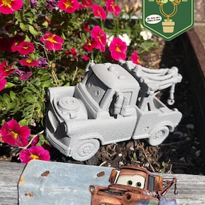 May include: A gray concrete Mater tow truck statue sits in a planter with vibrant pink flowers. A colorful cartoon version of Mater is also present. The planter has a metal edge and is filled with mulch.