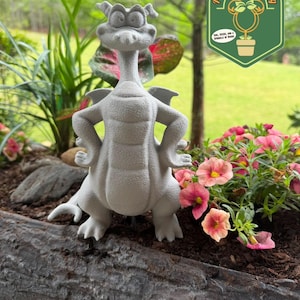 May include: A gray concrete dragon statue with its arms crossed, wings, and a long neck, placed in a garden bed with pink flowers and greenery. The statue is in front of a green background with a logo that says "Pixie Dust Gardening".