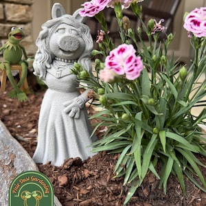 May include: A gray concrete Miss Piggy statue stands in a planter with pink and white flowers. A Kermit the Frog figure sits nearby. The planter is filled with brown mulch, and the logo for Pixie Dust Gardening is visible.