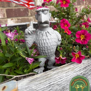 May include: A gray concrete garden statue of a character with a long nose and raised finger, surrounded by pink and red flowers. The statue is in a wooden planter box. An American flag is in the background.