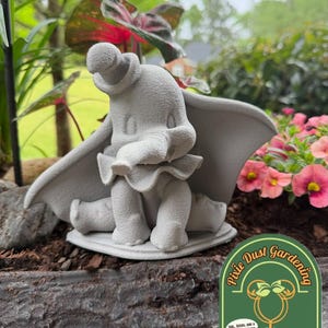May include: A gray concrete statue of Dumbo the elephant, seated with his ears spread. The statue is in a garden setting, surrounded by plants and flowers. The statue is on a base, and the image includes a logo for "Pixie Dust Gardening."