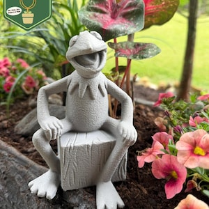 May include: A gray frog statue sitting on a tree stump, surrounded by plants and flowers. The frog has a smiling expression and is wearing a collar. The image includes a green and gold logo that says "Pixie Dust Gardening".