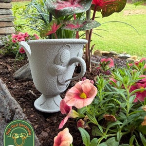 May include: A gray, textured teacup-shaped planter with a handle and a decorative face. The planter is surrounded by pink and red flowers and lush green plants. The background features a green lawn and a stone wall.