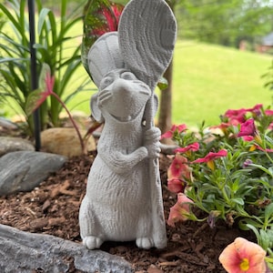 May include: A gray concrete statue of a mouse holding a large spoon. The mouse is smiling and standing upright. The statue is placed in a garden bed with flowers and greenery in the background.