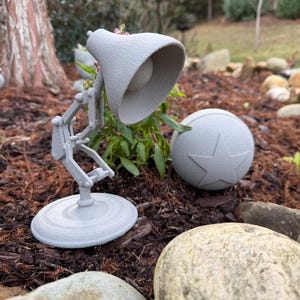 May include: A light gray desk lamp with a ball featuring a star design. The lamp has a round base and adjustable arm. The ball is next to the lamp. The objects are outdoors.