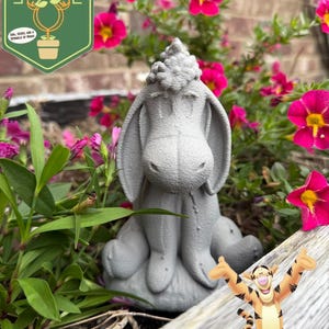 May include: A gray concrete statue of Eeyore from Winnie the Pooh, sitting among pink flowers and greenery. The statue is in a garden setting, with a cartoon image of Tigger in the lower right corner. The image also includes a logo that says "Pixie Dust Gardening".