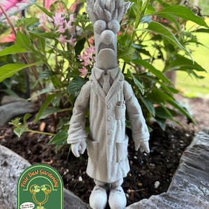 May include: A light gray 3D printed figurine of a scientist character with wild hair, a lab coat, a tie, and large eyes. The figure stands upright with a wide-eyed expression. A green logo with the text Pixie Dust Gardening is in the corner.