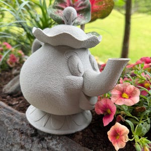 May include: A gray, sculpted teapot with a smiling face, spout, and a decorative lid. The teapot is sitting in a planter box with pink and yellow flowers and green foliage in the background.