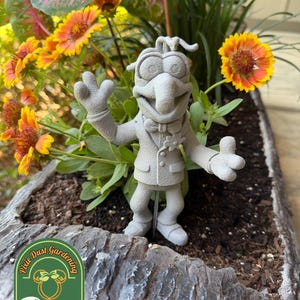 May include: A gray, textured, 3D-printed figure of a character with a long nose, wearing a suit and bowtie, with one hand raised in a wave and the other extended. The figure is mounted on a thin metal stake and features a small flower on the lapel.