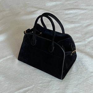 May include: A black suede handbag with two rounded handles and a structured design. The bag has a rectangular base and a slightly curved top, with a gold-colored clasp detail. The interior is visible and appears to be lined with a dark fabric.