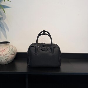 May include: A black leather handbag with a pebbled texture, featuring two rounded top handles, a central zipper closure with two matching leather pull tabs, and a structured rectangular base with rounded corners.