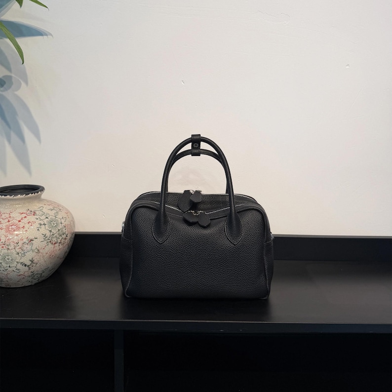 May include: A black leather handbag with a pebbled texture, featuring two rounded top handles, a central zipper closure with two matching leather pull tabs, and a structured rectangular base with rounded corners.
