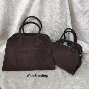 May include: Two brown suede handbags with rounded handles. The larger bag is rectangular, and the smaller bag is trapezoidal. Both bags have a soft, textured appearance and are set against a light-colored background. The text "With Branding" is at the bottom.