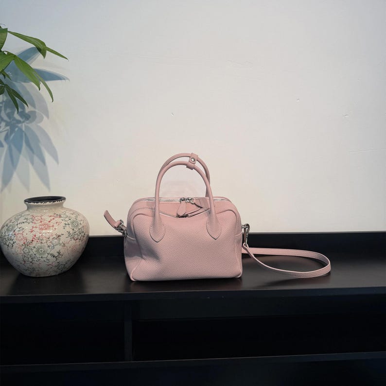 May include: A light pink leather handbag with a textured surface, featuring two top handles, a central zipper closure, and a detachable shoulder strap with silver-toned metal hardware. The bag has a structured, rounded rectangular shape.