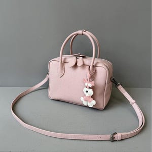 May include: A light pink handbag with a top handle and a detachable shoulder strap. The bag features a decorative dog charm and silver-tone hardware. The bag is made of a textured material.