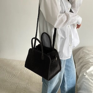 May include: A black handbag with a structured design, featuring two top handles and a long shoulder strap. The bag has a rectangular shape with rounded edges and a small gold-colored clasp. The bag is made of a soft, velvety material.