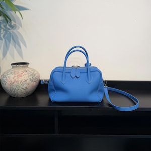 May include: A bright blue leather handbag with a textured surface, featuring two top handles, a silver-toned double zipper closure, and a detachable shoulder strap. The bag has a structured, rectangular shape with rounded corners.