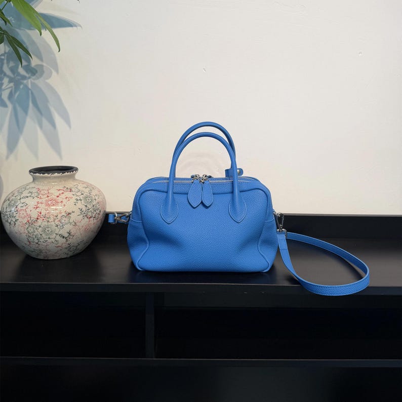 May include: A bright blue leather handbag with a textured surface, featuring two top handles, a silver-toned double zipper closure, and a detachable shoulder strap. The bag has a structured, rectangular shape with rounded corners.