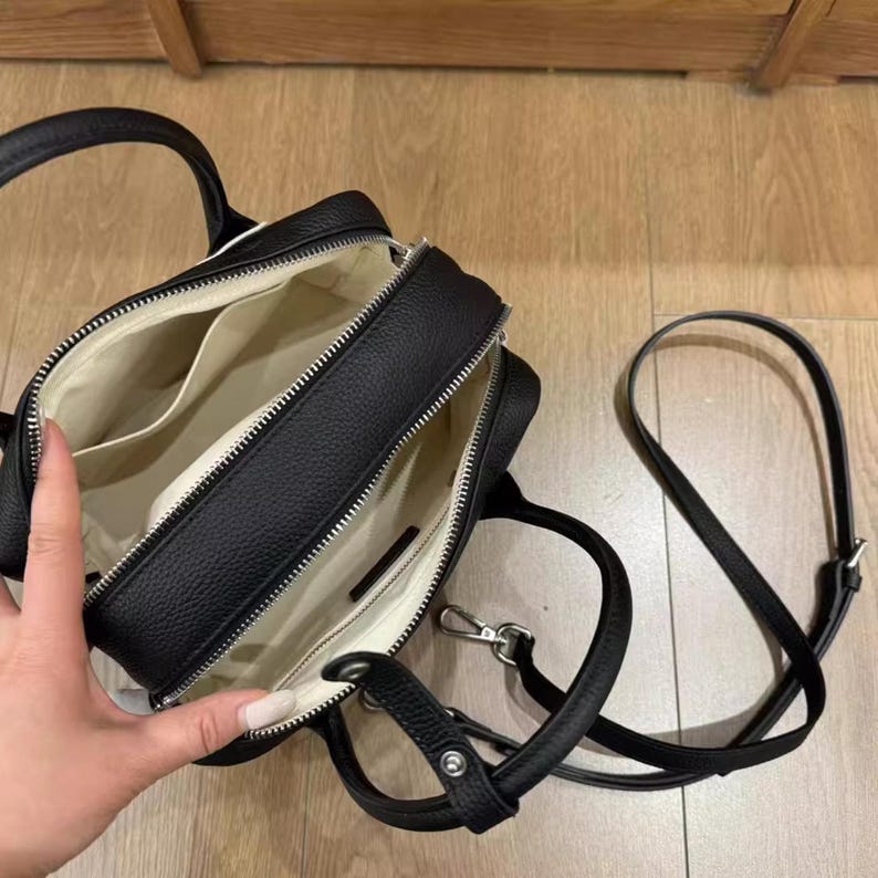 May include: Black, open handbag with a top handle and a detachable shoulder strap. The bag has a zippered closure and a light-colored interior with multiple pockets. The exterior is textured.