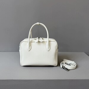 May include: A white, rectangular handbag with a textured surface, two short handles, and a detachable shoulder strap. The bag has a zipper closure and silver-tone hardware. The strap is approximately 40 inches long.