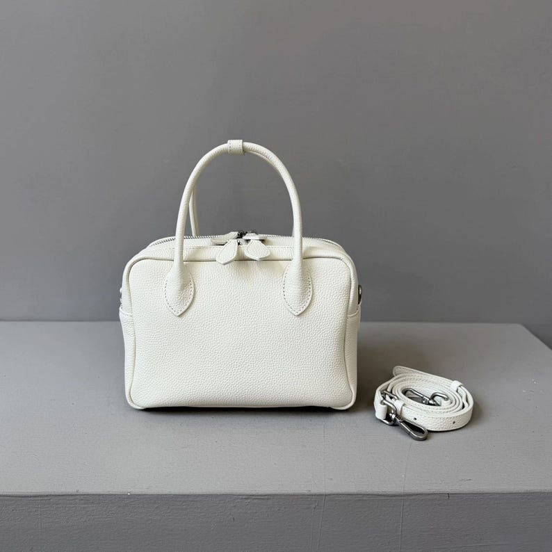 May include: A white, rectangular handbag with a textured surface, two short handles, and a detachable shoulder strap. The bag has a zipper closure and silver-tone hardware. The strap is approximately 40 inches long.
