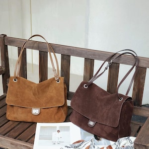 Könnte beinhalten: Zwei Schultertaschen aus Wildleder, eine in Hellbraun und eine in Dunkelbraun, jeweils mit rechteckiger Klappe, silberfarbenem Drehverschluss aus Metall und zwei dünnen Schulterriemen, die mit silberfarbenen Metallösen am oberen Rand befestigt sind.