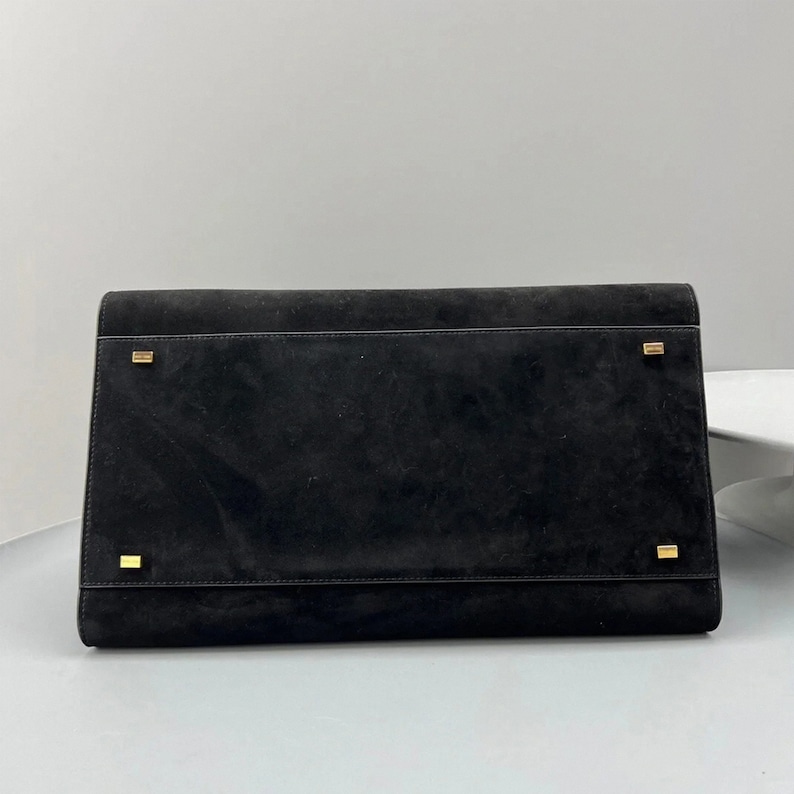 May include: A black suede clutch with a rectangular shape. The clutch features gold-toned metal accents on the front and a flap closure. The bag is displayed against a neutral background.