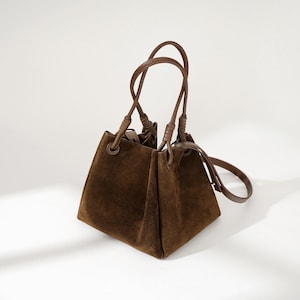 May include: A brown suede tote bag with knotted leather handles and a long strap. The bag has a square shape and a slightly slouchy look.
