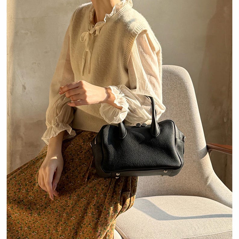 May include: A black leather handbag with a pebbled texture, two top handles, and silver-tone metal feet on the base. The bag features a rounded rectangular shape with a zippered closure along the top edge and visible stitching details.