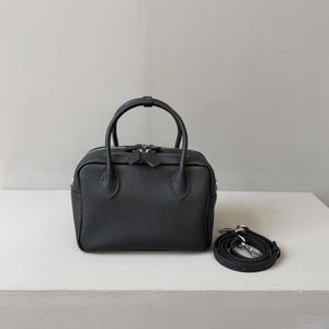 May include: A black, textured leather handbag with a rectangular shape, two top handles, and a top zipper closure. The bag features silver-tone hardware and includes a detachable, adjustable shoulder strap coiled beside it.