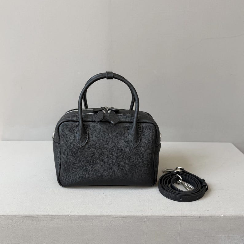 May include: A black, textured leather handbag with a rectangular shape, two top handles, and a top zipper closure. The bag features silver-tone hardware and includes a detachable, adjustable shoulder strap coiled beside it.