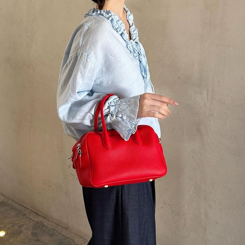 May include: A vibrant red leather handbag with a textured finish, featuring two sturdy top handles, a zippered closure, and silver-toned hardware. The bag has a structured rectangular shape with rounded corners and small metal feet on the base.