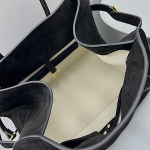 May include: Black suede tote bag with a structured design. The bag features a spacious interior with a light-colored lining. Gold-toned hardware accents the straps and closures, adding a touch of elegance to the overall design.