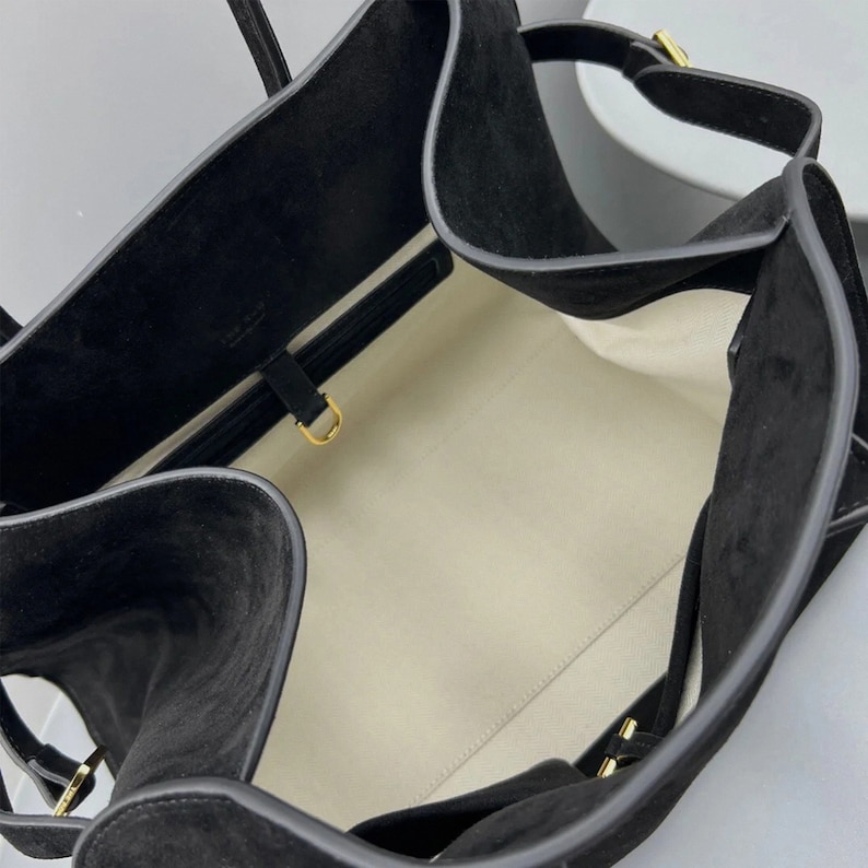 May include: Black suede tote bag with a structured design. The bag features a spacious interior with a light-colored lining. Gold-toned hardware accents the straps and closures, adding a touch of elegance to the overall design.