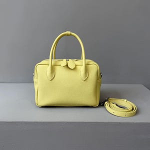 May include: A pale yellow handbag with a textured surface. The bag has two short handles and a detachable shoulder strap. The bag has a rectangular shape with a zipper closure and a small pocket on each side.