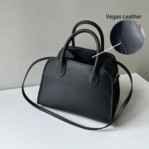 May include: Black vegan leather handbag with two top handles and a detachable shoulder strap. The bag has a structured shape with a stitched design detail. The image includes a close-up showing the texture of the vegan leather.