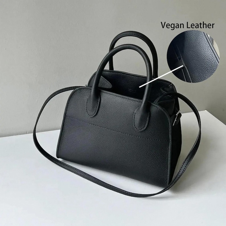 May include: Black vegan leather handbag with two top handles and a detachable shoulder strap. The bag has a structured shape with a stitched design detail. The image includes a close-up showing the texture of the vegan leather.
