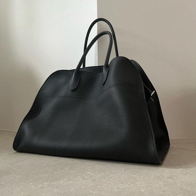 May include: A large, black leather tote bag with two curved handles. The bag has a textured surface and a spacious design, suitable for carrying various items. The bag is set against a neutral background, highlighting its sleek and minimalist aesthetic.