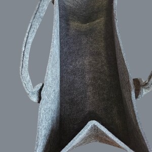 May include: A gray felt tote bag with two handles. The bag has a rectangular shape with a V-shaped bottom. The interior of the bag is visible, showing the felt texture. The handles are attached to the top of the bag.