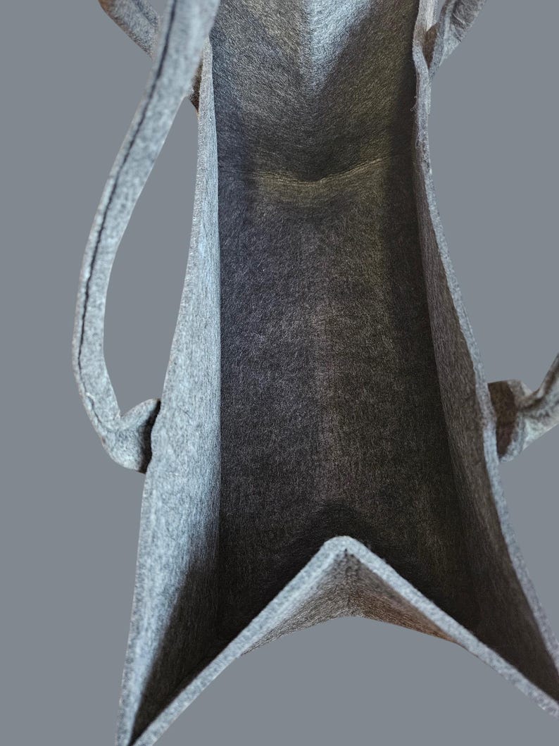 May include: A gray felt tote bag with two handles. The bag has a rectangular shape with a V-shaped bottom. The interior of the bag is visible, showing the felt texture. The handles are attached to the top of the bag.