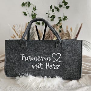Puede incluir: Una bolsa de mano de fieltro gris con texto blanco que dice "Trainerin mit Herz" y un símbolo de corazón blanco.
