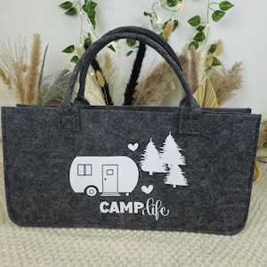 May include: A gray felt tote bag with two handles. The bag features a white graphic of a camper, two trees, and the words "CAMP life". The bag is rectangular and sits on a cream-colored surface.