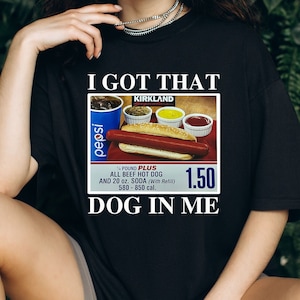 May include: Black t-shirt with a white graphic of a hot dog, soda, and condiments. The graphic includes the text "I got that", "Kirkland Signature", "Pepsi", "¼ pound plus", "All beef hot dog", "and 20 oz. soda (with refill)", "580-850 cal.", and "1.50". The text "Dog in me" is below the graphic.