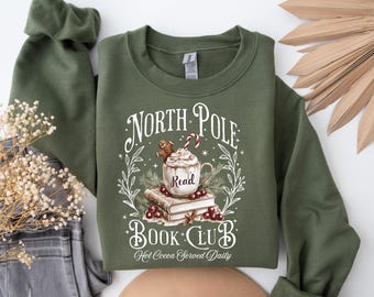 May include: Green long-sleeve sweatshirt with a white graphic design of a mug of hot cocoa with a gingerbread man and the text "North Pole Book Club. Hot Cocoa Served Daily."