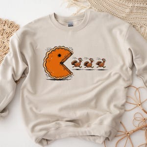 May include: Beige sweatshirt featuring a humorous graphic of a pie-shaped Pac-Man chasing three turkeys. The pie is orange with a brown crust, and the turkeys are brown. The design is a playful take on Thanksgiving.