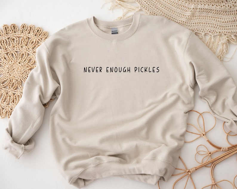 K&ouml;nnte beinhalten: Ein hellbeiger Pullover mit dem Aufdruck "NEVER ENOUGH PICKLES" in Schwarz auf der Vorderseite.
