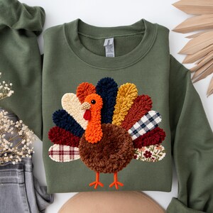 May include: Olive green sweatshirt featuring a colorful turkey design. The turkey has a brown pom-pom body, orange head, and a vibrant tail with various patterned and textured elements. The design is centered on the front of the long-sleeved top.