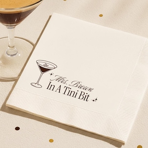 Può includere: Tovaglioli da cocktail color crema con un bicchiere da martini stampato e la scritta "Mrs. Brown In A Tini Bit". I tovaglioli sono piegati e hanno un bordo strutturato. Un bicchiere da martini con una bevanda marrone scuro e chicchi di caffè è sullo sfondo.